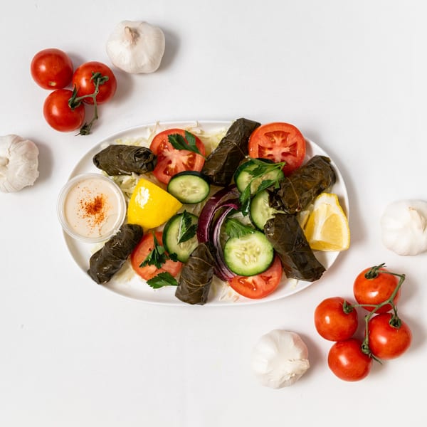 Middle-eastern dolmades with fresh salad, hummus, and lemon on a white plate, surrounded by tomatoes and garlic.