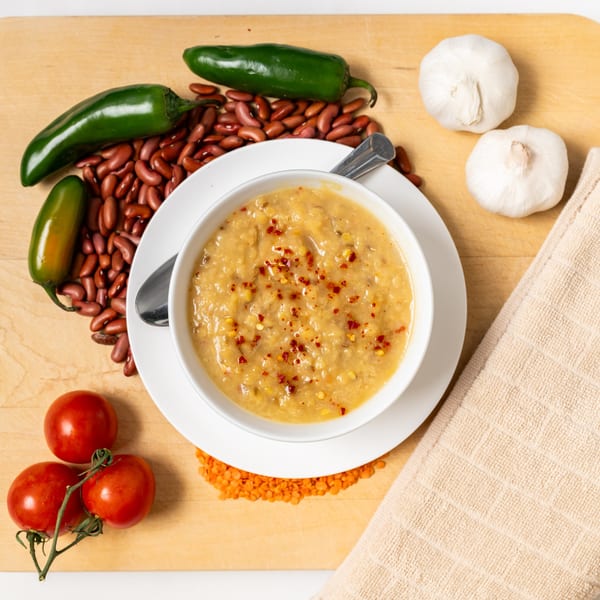 A bowl of Pita Pocket Eatery's lentil soup with chili flakes, surrounded by beans, peppers, garlic, and tomatoes.