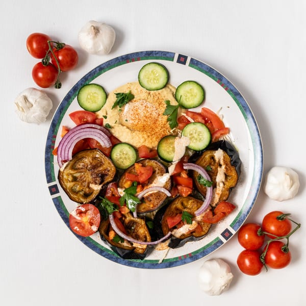 Middle-eastern dish with grilled eggplant, hummus, tomatoes, cucumbers, and red onions on a plate.